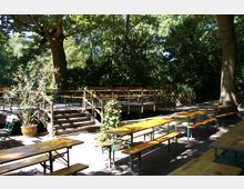 Ein Biergarten im Freien unter großen Bäumen mit Holzbänken und Tischen auf Kiesboden. In der Mitte befindet sich eine erhöhte Holzplattform mit Geländer, erreichbar über Treppen, umgeben von Pflanzen und grünem Laubwerk.