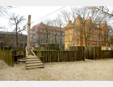 Ein Spielplatz mit Holzkonstruktionen auf sandigem Boden, umgeben von kahlen Bäumen. Im Hintergrund stehen historische Backsteingebäude mit roten Ziegeldächern.