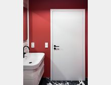 Badezimmer mit einer roten Wand, einer weißen Tür mit schwarzem Türgriff, einem weißen Waschbecken mit schwarzer Armatur auf der linken Seite und einem Boden mit schwarz-weißem Marmormuster.