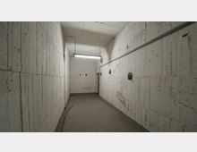 Schmaler, rechteckiger Raum mit rohen Betonwänden, einer abgenutzten Betonbodenfläche und sichtbaren Leitungen an der Decke und der Wand. Eine Leuchtstofflampe hängt zentral von der Decke, und an der rechten Wand sind runde technische Vorrichtungen angebracht.