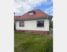 Seitenansicht eines eingeschossigen Wohnhauses mit weiß verputzter Fassade, rotem Ziegeldach und kleinem Dachgaubenfenster. Vor dem Haus liegt eine Rasenfläche; sichtbar sind zwei Fenster, eine Regenrinne und ein Sockel aus rotem Backstein.