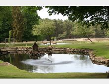 Ein öffentlicher Park mit einem kleinen Teich im Vordergrund, von Steinen umgeben, und einem Holzhäuschen am Teichrand. Im Hintergrund grasen zwei Hirsche auf einer Wiese mit Bäumen und einem Wald als Kulisse.