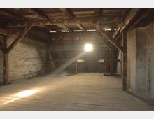 Ein leerer Dachboden mit sichtbaren Holzbalken und Holzboden; an der Stirnwand fällt durch ein kleines Fenster ein Lichtstrahl ins Innere. Die Wände sind aus unverputztem Stein und das Dach besteht aus Holzlatten.