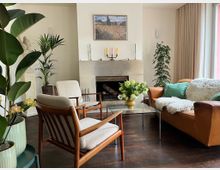 Helles Wohnzimmer mit zentralem, weiß verkleidetem Kamin und einem Landschaftsbild darüber, davor ein Glas-Couchtisch mit Blumenstrauß. Rechts steht ein braunes Ledersofa mit Kissen und Decke neben bodentiefen Fenstern mit Vorhängen; links sind zwei Holzsessel und mehrere große Zimmerpflanzen auf dunklem Holzboden angeordnet.