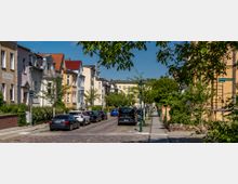 Straßenszene in einem Wohngebiet mit gepflasterter Fahrbahn, geparkten Autos und mehrstöckigen Altbauhäusern mit hell gestrichenen Fassaden. Links und rechts der Straße stehen Bäume und Laternen, und ein Straßenschild zeigt „Danckelmannstraße“.