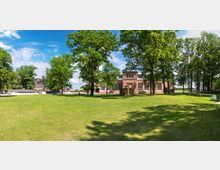 Grüne Parkanlage mit großen, schattenspendenden Bäumen und einer historischen Backsteinfassade im Hintergrund, die zu einem langgestreckten Gebäude mit großen Fenstern gehört. Der Himmel ist klar mit vereinzelten Wolken, und Wiesenflächen dominieren den Vordergrund.