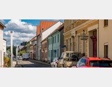 Eine schmale Kopfsteinpflasterstraße mit farbigen, historischen Wohnhäusern und Schildern, darunter "Zum Kommandanten". Am Straßenrand parken Autos, und im Hintergrund sind Bäume und ein Kirchturm zu sehen.