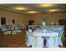 Großer Festsaal mit Holzboden, gedeckten Rundtischen und Stühlen, die mit weißen Hussen und grün-transparenten Schleifen dekoriert sind. Luftballons und Tischdekorationen in Grün und Weiß schaffen eine festliche Atmosphäre, während moderne, runde Deckenleuchten den Raum beleuchten.