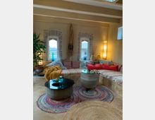 Gemütlicher Wohnbereich mit einer großen, bodenintegrierten Eckbank, die mit bunten Kissen und gemusterten Decken dekoriert ist. Auf dem beigen Boden liegen runde Teppiche in Natur- und bunten Farben; zwei runde, geflochtene Tische mit Glasplatte stehen im Zentrum, umgeben von Pflanzen und sanfter Beleuchtung.