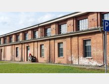 Ziegelsteinfassade eines einstöckigen Gebäudes mit großen rechteckigen Fenstern, teilweise über zwei Ebenen. Im Vordergrund befinden sich eine Grünfläche, ein Gehweg und ein Schild mit einem "P"-Symbol.