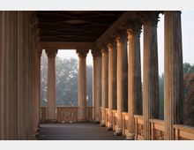 Eine überdachte Säulengalerie mit korinthischen Säulen, die auf beiden Seiten symmetrisch angeordnet sind. Die Szenerie wird von weichem, seitlichem Sonnenlicht beleuchtet, im Hintergrund sind Bäume im Dunst zu erkennen.