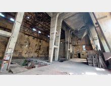 Innenansicht einer verlassenen Industriehalle mit hohen Betonsäulen, rostigen Metallstrukturen und sichtbarem Mauerwerk. Die Decke ist teilweise beschädigt, und am Boden sind Vertiefungen sowie verstreute Materialien sichtbar.