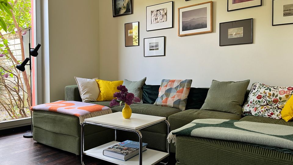 Gemütliches Wohnzimmer mit grünem Ecksofa, bunten Kissen und Decken sowie einem weißen Couchtisch mit Vase; an der Wand hängt eine Galerie aus gerahmten Bildern. Links sorgt eine große Fensterfront mit Blick ins Grün und eine Stehlampe für viel Tageslicht.