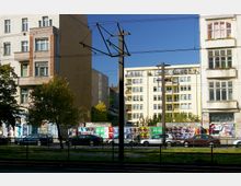 Straßenszene mit mehrstöckigen Wohngebäuden, teils mit Balkonen, und einer mit Plakaten bedeckten Mauer im Vordergrund. Autos parken entlang der Straße, und Gleise sowie Oberleitungsmaste einer Straßenbahn sind sichtbar.