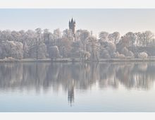 Ein ruhiger See spiegelt eine Uferlandschaft mit kahlen, von Raureif bedeckten Bäumen und einem hohen Turm mit spitzer Ziegelarchitektur im Hintergrund wider. Der Himmel ist hell und neblig, was der Szenerie eine weiche, frostige Stimmung verleiht.