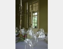 Eleganter, festlich gedeckter Tisch mit weißen Stoffservietten, Gläsern und einem silberfarbenen Kerzenleuchter im Vordergrund. Im Hintergrund sind geöffnete Doppelfenster mit Blick nach draußen sichtbar.