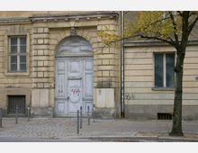 Fassade eines historischen Gebäudes mit vergilbtem Putz und dekorativem, verwittertem Holztor in einem Steinbogen. Links und rechts sind Fenster zu sehen, davor führt ein gepflasterter Gehweg mit Pollern entlang; ein Baum steht rechts im Bild.
