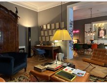Wohnzimmer mit grauen Wänden, Stuckdecke und Holzboden, eingerichtet mit großem Holzschrank, Sesseln, Lederhockern und Couchtisch unter einer großen Hängelampe. Im Hintergrund führt eine breite Türöffnung in ein Esszimmer mit langem Tisch, bunten Stühlen und einem Geweih-Kronleuchter.