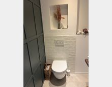 Moderne Gästetoilette mit einer wandhängenden Toilette vor einer Wand mit weißen Fliesen im U-Bahn-Stil. Links steht ein geflochtener Korb für Zeitschriften, oben in einer Wandnische eine Vase mit getrockneten Gräsern, daneben aufgerollte Handtücher.