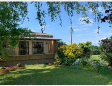 Ein flaches Wohngebäude mit großen Fenstern, umgeben von einem Garten mit grünem Rasen, bunten Blumenbeeten und Büschen. Im Hintergrund sind eine offene Landschaft und ein blauer Himmel zu sehen.