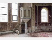 Innenansicht einer alten, verwitterten Kirche mit einer weiß bemalten Kanzel auf einem dekorierten Sockel. Die Wände sind teilweise abgeblättert, mit verblassten Mustern und Graffiti, flankiert von hohen Fenstern mit milchigem Glas, und ein Teppich bedeckt den gemusterten Steinboden.