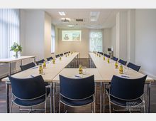 Konferenzraum mit U-förmig angeordneten weißen Tischen und dunkelblauen Stühlen, ausgestattet mit Getränken, Gläsern und Schreibutensilien. Im Hintergrund befinden sich große Fenster mit weißen Jalousien sowie ein Projektor und eine Leinwand.