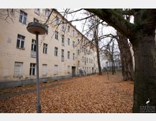 Hof eines alten Wohngebäudes mit beige verputzter Fassade, mehreren Fenstern und geschlossenen Fensterläden. Der Boden ist mit Herbstlaub bedeckt, Bäume und Laternen stehen im Hof.