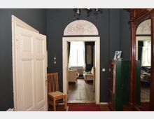 Flur eines historischen Hauses mit dunklen Wänden, einer offenen Tür zum angrenzenden Wohnzimmer und verziertem Bogengiebel über dem Türrahmen. Im Vordergrund befinden sich ein Holzstuhl, ein antiker Schrank mit Glasfront und ein Spiegel.