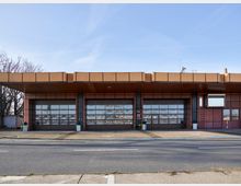 Flach gehaltenes Gebäude mit großen, verglasten Sektionaltoren, vermutlich eine ehemalige Garage oder Werkstatt. Das Gebäude hat eine orange-braune Fassade, ein überstehendes Flachdach und befindet sich an einer Straße; im Hintergrund sind kahle Bäume sichtbar.