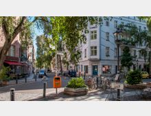 Straßenszene in einer ruhigen Wohngegend mit begrünten Gehwegen; Eckgebäude mit mehreren Stockwerken, einer Ladenfront im Erdgeschoss und Balkonen. Fahrräder sind an Ständern abgestellt, ein Mülleimer und Straßenschilder sind im Vordergrund sichtbar, während Bäume Schatten spenden.