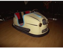 Ein gelbes, retro-stilisiertes Autoscooter-Fahrzeug mit blauen und schwarzen Akzenten sowie roten Polstersitzen, ausgestellt auf einem Holzfußboden in einem dunklen Raum.