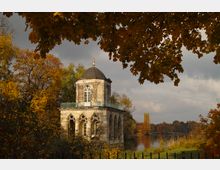 Kleiner achteckiger Pavillon aus Stein mit Kuppeldach und gotischen Rundbogenfenstern, umgeben von herbstlich gefärbtem Laub und angrenzendem See im Hintergrund. Ein Ast mit goldbraunen Blättern ragt im Vordergrund ins Bild.