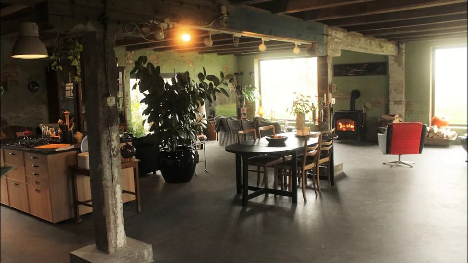 Offener Wohnraum in einem industriellen Stil mit sichtbaren Holzbalken und Betonboden. Die Einrichtung umfasst eine Küchenzeile, einen Esstisch mit Stühlen, große Zimmerpflanzen, einen gemütlichen Holzofen und große Fenster, die Tageslicht einlassen.