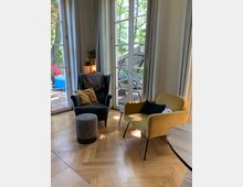 Ein gemütlicher Wohnbereich mit einem dunklen Sessel, passendem Hocker und einem gelben Sessel, beide vor bodentiefen Fenstern mit weißen Vorhängen. Draußen ist eine Terrasse mit Gartenmöbeln und Bäumen sichtbar, der Boden ist aus Holzparkett im Fischgrätmuster.