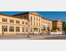 Gelbes, zweistöckiges Bahnhofsgebäude aus der Gründerzeit mit symmetrischen Fenstern und einem Dreiecksgiebel. Vor der Fassade befinden sich eine Bushaltestelle, Laternen und junge Bäume auf einem gepflasterten Vorplatz.