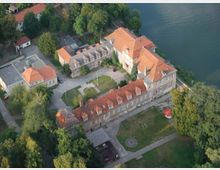 Luftaufnahme eines historischen Gebäudekomplexes mit roten Ziegeldächern, umgeben von Bäumen. Der Komplex liegt in Wassernähe und umfasst einen Innenhof mit Rasenflächen und Wegen.