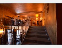 Innenraum eines zweistöckigen Restaurants in warmem, goldenen Licht. Im Vordergrund führt eine Treppe mit dekorativem Geländer zur oberen Etage mit Tischen, Stühlen und beleuchteten Wandleuchten; im unteren Bereich sind Tische und große Fenster erkennbar.