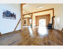 Weitläufiger Innenraum mit Holzboden, sichtbaren Holzbalken und einer Ziegelwand mit eingebogenem Fenster. Im Raum befinden sich ein Kaminofen, eine weiße Tür sowie eine Fotografie an der weißen Wand.