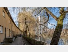 Ein gepflasterter Weg führt entlang einer Reihe beige gestrichener Reihenhäuser mit kleinen Treppen vor den Eingängen. Rechts des Weges stehen Weidenbäume an einem Flussufer, im Hintergrund ist ein mehrstöckiges Wohngebäude zu sehen.