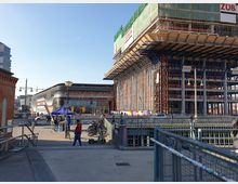 Straßen- und Platzszene mit einer modernen, schwarzen Gebäudefassade der "East Side Mall" links und einem angrenzenden großen Baustellengerüst eines Hochbaus rechts. Im Vordergrund befinden sich Fußgänger, Fahrräder und ein Elektro-Scooter, umgeben von Geländern und Baustellenabsperrungen.