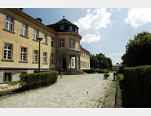 Historisches Gebäude mit gelber Fassade und weißen Fensterrahmen, sowie einem runden Eingangsbereich mit Säulen. Der gepflasterte Vorplatz ist von Grünflächen und Hecken umgeben, im Hintergrund sind einige Bäume zu sehen.