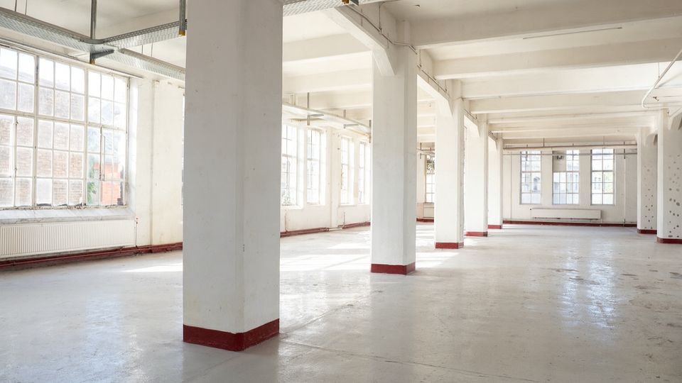 Großer, leerer Industrie- oder Loftinnenraum mit weißen Stützpfeilern, Betonboden und hoher Decke mit sichtbaren Leitungen und Trägern. Entlang der Außenwände befinden sich große, mehrteilige Fenster, durch die viel Tageslicht einfällt.