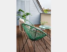 Kleine Dachterrasse mit Holzboden, darauf zwei grüne Stühle mit geschnürter Rückenlehne und ein passender kleiner Tisch. Im Hintergrund eine hellgraue Holzverkleidung mit Fensterläden sowie Topfpflanzen als Dekoration.