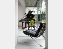 Modern minimalist living room with a black grand piano and a black leather sling chair on a light floor, beside floor-to-ceiling glass walls. A small table with yellow tulips sits near the window, looking out to a garden with trees and a wooden fence.