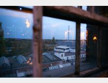 Blick durch ein mehrteiliges Fenster auf ein Industriegelände mit Gebäuden in Flachdachbauweise und runden Dachfenstern. Im Hintergrund sind Bäume und ein Schornstein bei Dämmerung zu sehen, mit Spiegelungen von Innenbeleuchtung auf der Fensterscheibe.
