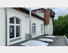 Außenansicht eines Gebäudes mit heller Fassade und roten Dachziegeln, das große, weiße Fenster mit Rundbogen aufweist. Im Vordergrund ist ein Flachdach mit mehreren Oberlichtern und einem Metallrohr zu sehen, im Hintergrund sind Bäume sichtbar.