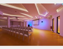 Ein moderner Veranstaltungsraum mit gelbem Boden, weißen Stühlen in Reihen und einer großen Leinwand mit der Aufschrift "Space Shack" an der Vorderwand. Die Decke hat dekorative, beleuchtete Elemente, und die Wände sind in neutralen Farben gehalten, unterstützt durch farbige LED-Beleuchtung.