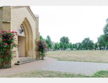 Backsteinfassade einer Kirche mit einem Spitzbogenportal, umrahmt von blühenden Rosensträuchern. Im Vordergrund befindet sich ein gepflasterter Weg, dahinter eine grüne Wiese, gesäumt von Bäumen und Wohnhäusern.