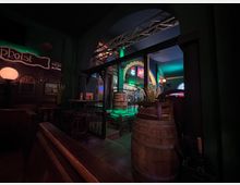 Innenraum einer dunklen Bar mit grüner Akzentbeleuchtung, Holzbänken und kleinen Tischen sowie mehreren großen Holzfässern im Vordergrund. Ein großer Rundbogen führt in einen blau beleuchteten Bereich mit Barwand, Bildern und Neonanzeigen.