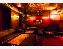 Gemütlicher Bar- oder Lounge-Innenraum mit holzvertäfelten Wänden, Ledersofas und niedrigen Holztischen mit kleinen Kerzen. Warme, gedämpfte Beleuchtung mit Deckenleuchte und Wandlampen, sichtbaren Lüftungsrohren sowie roter Akzentwand mit Spiegel.